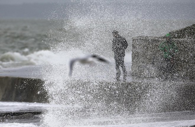Một người đàn ông đứng trước một cơn sóng vỗ ngay khi con mòng biển bay qua ở Portsmouth, Anh, ngày 13/11. Ảnh: AP.
