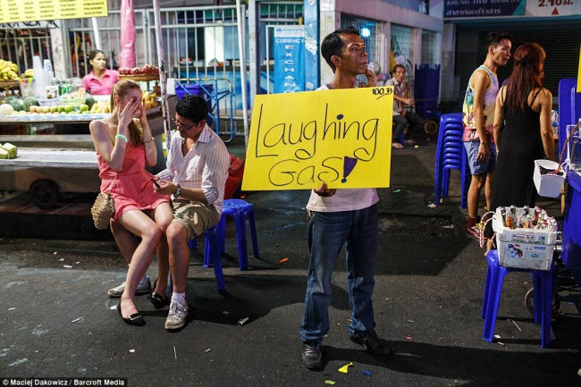 Cặp du khách nước ngoài ngồi nói chuyện với nhau, trong khi một người đàn ông cầm biển bán khí cười tại khu phố du lịch nhộn nhịp vào ban đêm ở Bangkok.