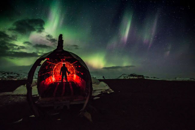 Người đàn ông khám phá một phần của chiếc máy bay bỏ hoang giữa phong cảnh mùa đông ở Iceland, khi ánh sáng bắc cực quang xuất hiện ở phía chân trời.